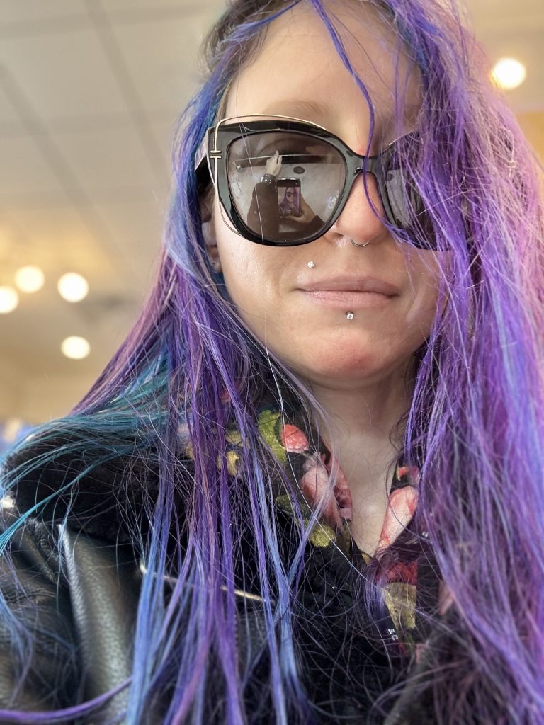 Photograph of Xexilia O. Shadows, 2025. She wears large, oversized sunglasses, a hoodie with a floral pattern taken from Renaissance paintings by Selkie, a deteriorating spiked rose faux-leather jacket over it by Blank NYC, and her hair is a combination of a blue-based purple, blue, and teal, and so long it reaches half-way down her spine. Xexilia's hair is parted on the left. Behind her are several recessed lights as she is at a nail salon. As she awkwardly smiles at the camera in a candid she took of herself, an uncommon choice of Xexilia due to her shyness, to send to her then-brand-new boyfriend of only a few days who had commented on missing her smile, so Xexilia sent this as her first attempt, taken while sipping a drink, lacked a smile when she took the photo to send to ease the loneliness they both felt. Once it was mentioned that seeing her smile was important, Xexilia tried again to capture a natural smile, but instead captured her awkward, social anxiety induced, somewhat humiliated, shy smile; Her boyfriend was happy enough with it, regardless, to become her fiancée. This image is on her blog archve of her professional art portfolio, with a link to a single post that explains what happened to her old blog, and how to access her new one.