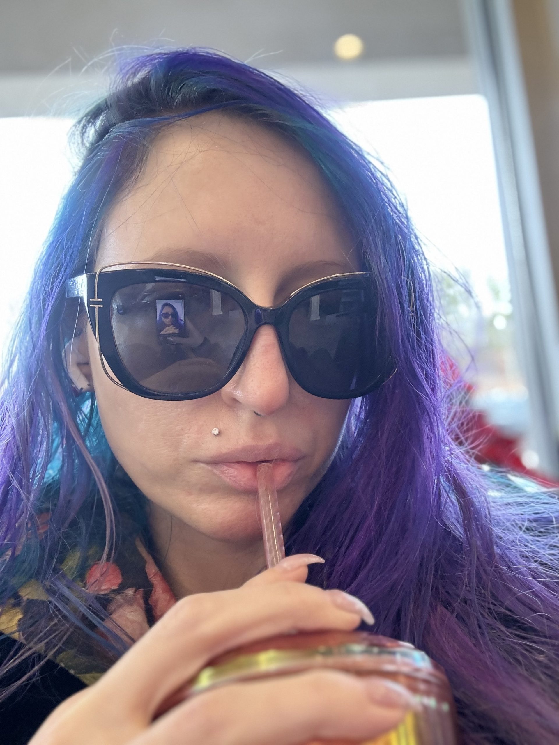 Photograph of Xexilia O. Shadows, 2025.  She wears large, oversized sunglasses, a hoodie with a floral pattern taken from Renaissance paintings by Selkie, a deteroiating spiked rose pleather jacket over it by Blank NYC, while drinking a homemade, sugar free, milk free, matcha frappe out of a large, orange and iridescent Starbucks purchased, personal cup she sips through a straw.  Behind her is a view to an overcast sky through a window and some distance bare tree branches, while just in front of the window, inside the nail salon, a corner of the red bloom of a poinsettia flower peers out behind Xexilia on the right of the viewer.  Only Xexilia herself remains in focus.  Her hair is a combination of a blue-based purple, blue, and teal, and so long it reaches half-way down her spine.  Xexilia's hair is parted on the left.  While excellent at photography, Xexilia hates taking 'selfies',  making this an extremely unusual photo of Xexilia due to her shyness, to send to her then beginning romance with her  polycule partner who remarked on wanting to see Xexilia smile again, so Xexilia sent this as her first attempt, taken while sipping a drink, lacking a  smile, as Xexilia was too afraid and shy to call more attention to herself by making it obvious she was taking a selfie. She tried again and despite the obvious embarrassment, her partner appreciated the effort in the end; Her partner was pleased enough, it would later turn out, to purpose.  Xexilia accepted.

The purpose of this image is art collectors, art lovers, curators, people hiring an artist, etc., supposedly want and expect to see a photo of the artist on their Biography page.  They also expect, people claim, a lot of other things to be much less informative or to include as many images as one would expect.